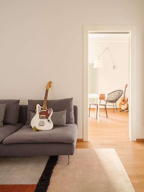 Minimalistic living room with grey sofa, white guitar, and open doorway revealing a chair.