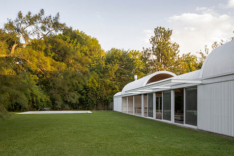 A modern, glass-enclosed home nestled in a lush, wooded setting with a large grassy lawn and pool.