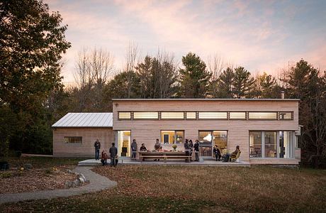 A modern, wooden building with large windows and a patio overlooking a forested landscape.