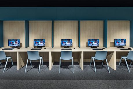 A modern computer room with sleek wooden desks, gray chairs, and multiple monitors.