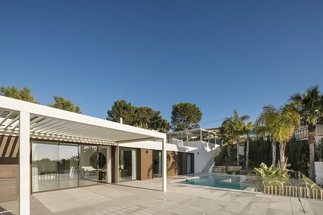 Modern white home with glass walls, pool, and lush landscaping under blue sky.