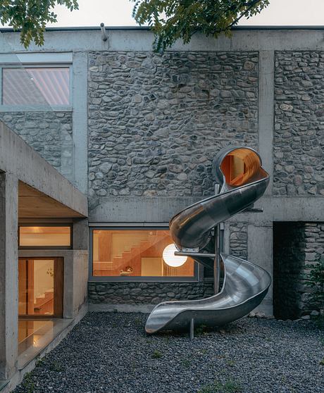 A modern, stone-clad house with a striking metal spiral slide leading to the entrance.