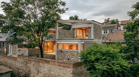 A modern stone and concrete building with large windows overlooking lush greenery.