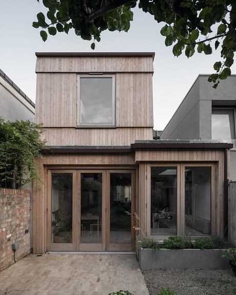 A modern, wooden-clad house with large windows and a paved entryway.