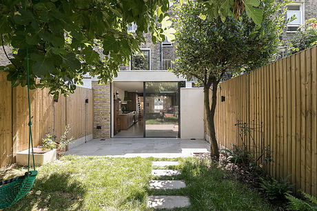 A modern glass-and-brick entryway opens to a lush backyard with paving stones and greenery.