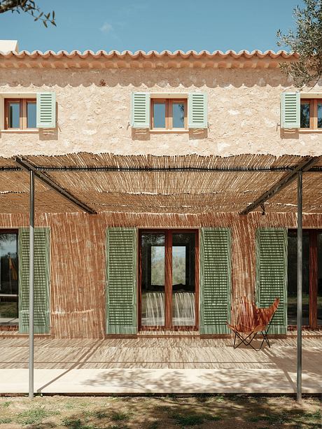 A rustic Mediterranean-style building with tiled roof, green shutters, and woven bamboo shade.