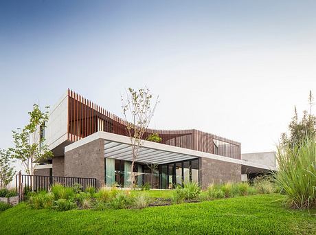 A modern architectural structure featuring a combination of concrete, wood, and glass elements, surrounded by a lush green landscape.