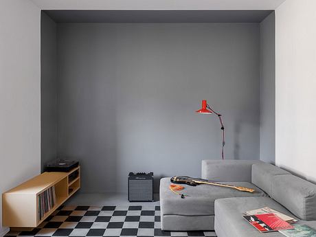 A minimalist living room with a gray sofa, a record player, and a red lamp on a checkered floor.