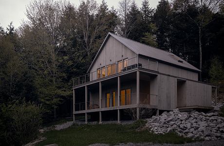 A modern cabin-style home with a wraparound porch and warm interior lighting.