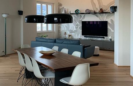 A spacious, modern living room with a wooden dining table, white chairs, and a stylish TV wall.