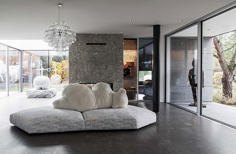 Large, modern living room with plush gray sofa, crystal chandelier, and glass walls overlooking nature.