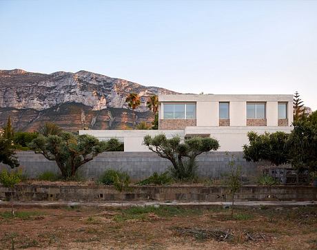 A modern, minimalist building with large windows nestled among desert vegetation and mountains.