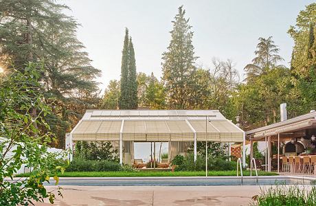 A modern, glass-enclosed poolside pavilion surrounded by lush greenery and trees.