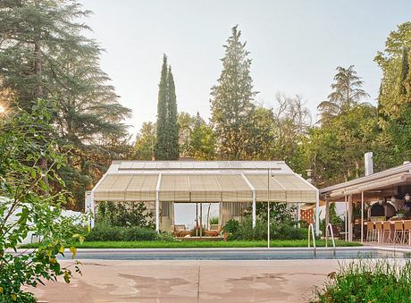 A modern, glass-enclosed poolside pavilion surrounded by lush greenery and trees.