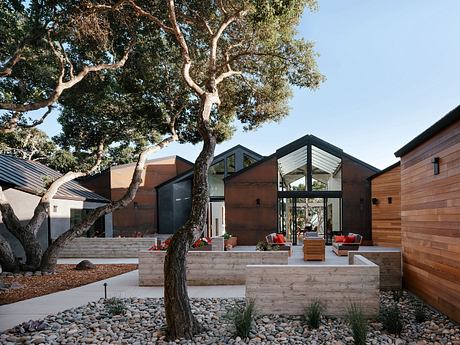 A modern, wooden-paneled home with a large tree and a paved outdoor patio area.