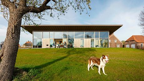 A modern glass-walled building with a wooden roof surrounded by a lush green lawn and an oak tree.