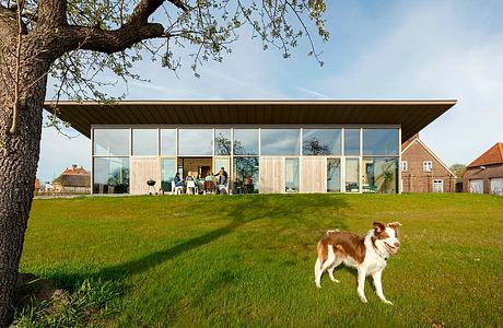 A modern glass-walled building with a wooden roof surrounded by a lush green lawn and an oak tree.