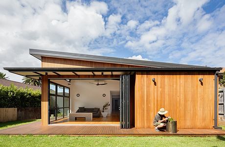 Modern wooden structure with covered patio and sliding glass doors, surrounded by lush greenery.