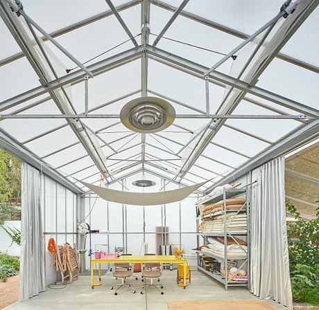 A modern greenhouse with a glass roof and metal frame, featuring a workspace with furniture.