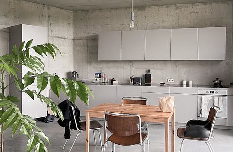 Minimalist kitchen and dining area with concrete walls, white cabinets, and a wooden table.