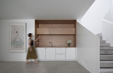 The image showcases a minimalist kitchen design with wooden panels, white cabinets, and a staircase leading upstairs.