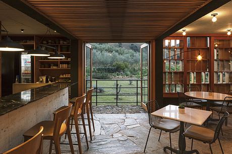 Rustic cabin interior with wooden beams, stone floors, and expansive window overlooking lush greenery.