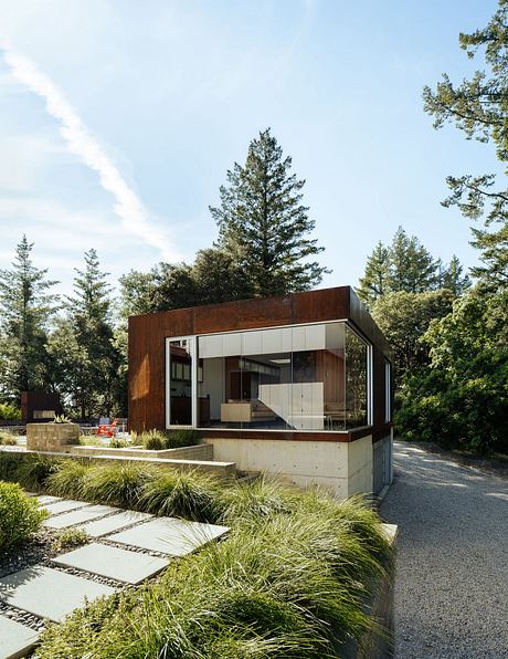 A modern, minimalist cabin surrounded by lush greenery and a gravel path leading to it.