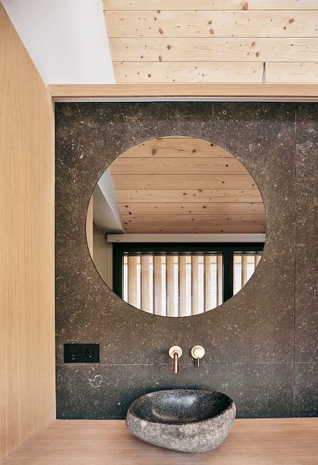 Circular mirror framed by wood-paneled ceiling, slate vanity with brass faucets.