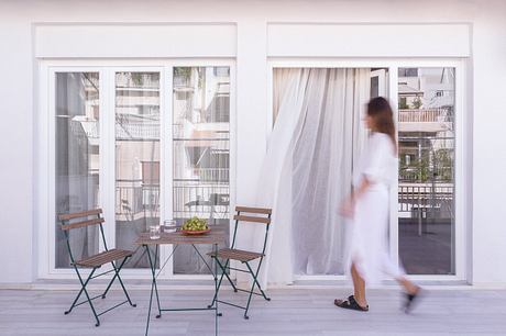 A bright, minimalist interior with large windows, white curtains, and a small dining area.