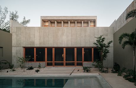 Minimalist concrete structure with wooden windows and doorways, surrounded by lush vegetation.