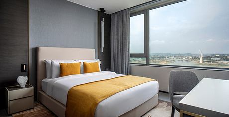 Modern hotel room with floor-to-ceiling windows, beige and gray color scheme, and yellow accent pillows.