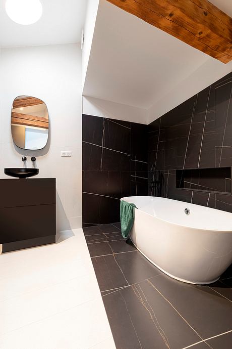 Modern bathroom with white freestanding tub, black tiled walls, and wooden ceiling beams.