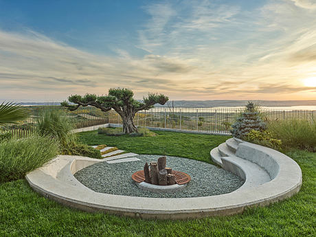 Landscaped backyard with concrete seating, fire pit, and lush greenery overlooking water.