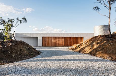 A modern, minimalist building with a large wooden door and a gravel driveway surrounded by trees.