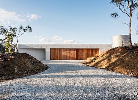 A modern, minimalist building with a large wooden door and a gravel driveway surrounded by trees.