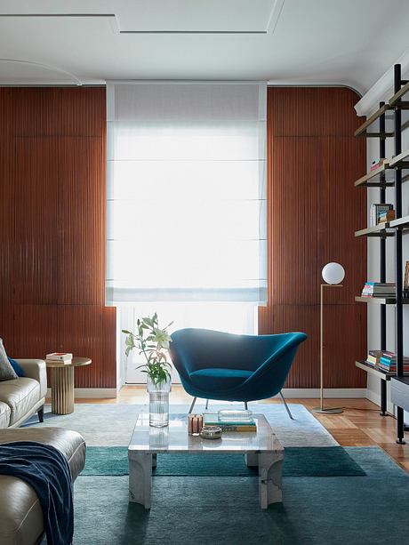 Warm-toned wooden paneling, plush blue chair, and sleek marble coffee table create a cozy ambiance.