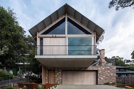 A modern two-story home with a pitched roof, exposed wooden beams, and a balcony overlooking greenery.