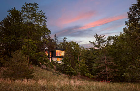 Modern, glass-walled house atop a wooded hilltop, framed by vibrant sunset sky.