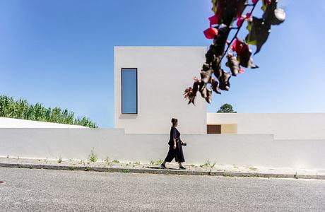 A modern, minimalist building with a single narrow window and vegetation surrounding it. A person in black clothing walking by.