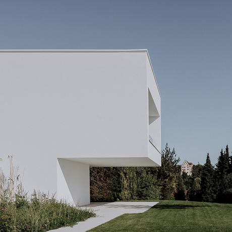A modern white structure with clean lines and a minimalist design, set against a forested backdrop.