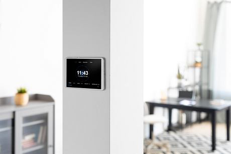 A modern digital clock mounted on a plain white wall, with a minimalist dining area in the background.