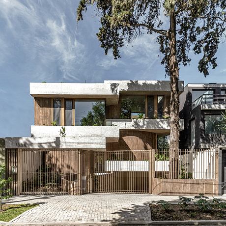 A modern, multi-level house with a mix of white concrete and warm wood finishes, surrounded by lush greenery.