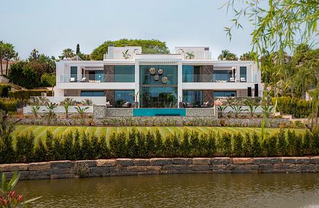 Modern multistory villa with lush landscaping, stone exterior, and balconies overlooking a pond.