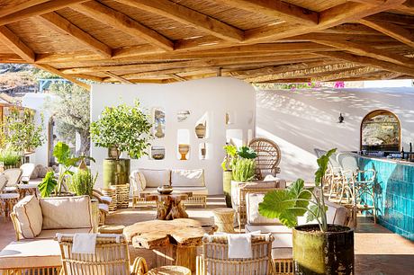 Rustic wooden beamed ceiling, white walls, rattan furniture, and lush greenery create an inviting outdoor living space.