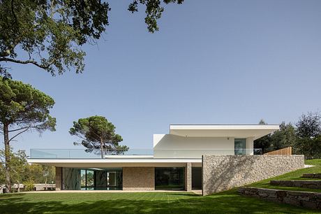 Modern two-story house with stone walls, glass, and sloping roof surrounded by lush greenery.