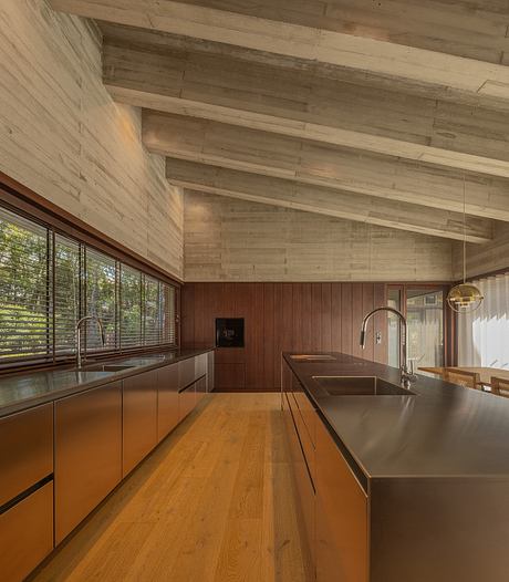 A modern open-plan kitchen with warm wood paneled walls and ceilings, stainless steel countertops, and large windows.