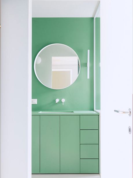 Minimalist bathroom with round mirror and green cabinetry against white walls.