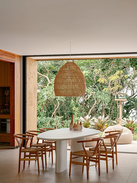 Cozy dining area with rattan pendant light, wooden chairs, and lush greenery views.