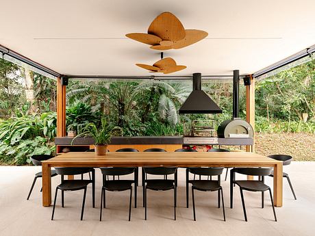 A modern, open-air dining area surrounded by lush greenery and featuring a wooden table, black chairs, and a unique fan-shaped light fixture.