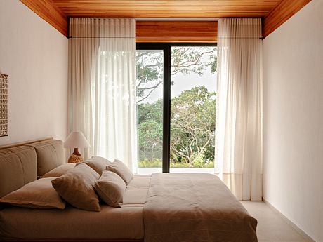 A cozy bedroom with a wooden ceiling, large windows overlooking a lush outdoor view, and plush textiles.